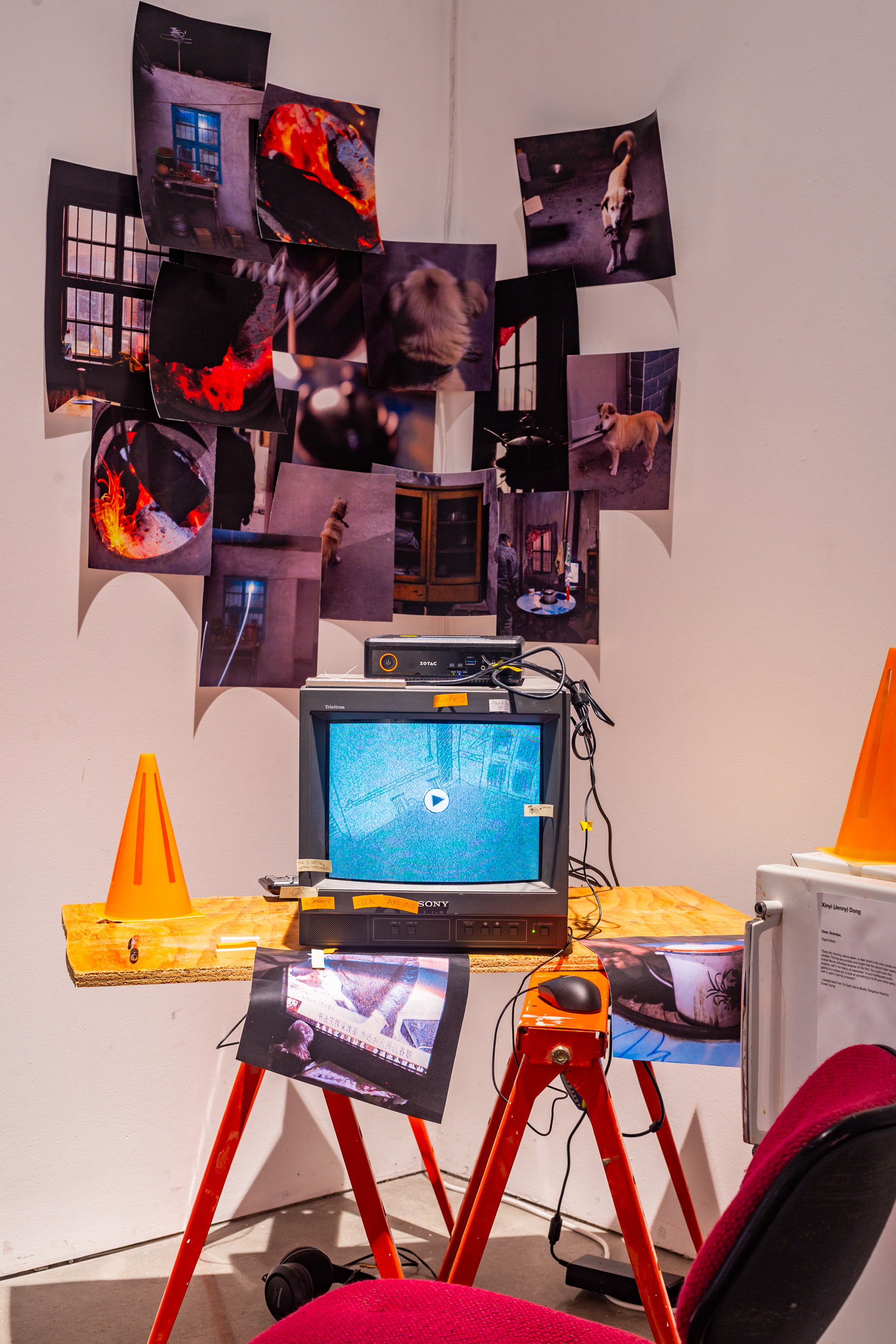 A sculptural piece, on the right is a white fridge with a plastic cone on top of it, in the center is a makeshift construction desk with plywood and an office chair, on top of it is a CRT tv with visuals on it, in the background is printed posters of photogrpahy.